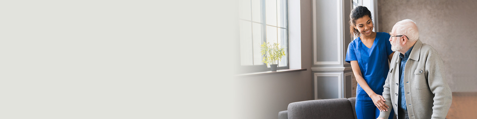 A nurse wearing blue scrubs standing next to an elderly gentleman next to a window