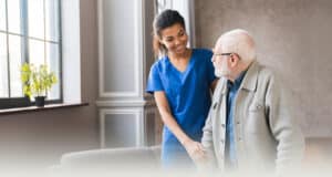 A nurse wearing blue scrubs standing next to an elderly gentleman next to a window